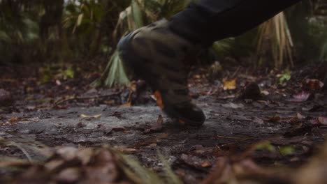 a close-up of a rugged hiking boot stepping on a muddy forest trail surrounded by fallen leaves and dense greenery. the scene evokes a sense of adventure, exploration, and connection with nature