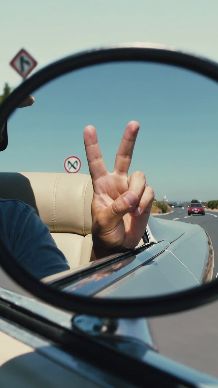 Vertical video: Reaching man's right hand making V sign in mirror as car driving on road for peace