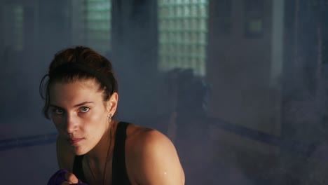 Portrait-of-a-young-martial-arts-female-athlete-shadowboxing-looking-in-the-camera-training-in-a-dark-room-with-smoke.-Her