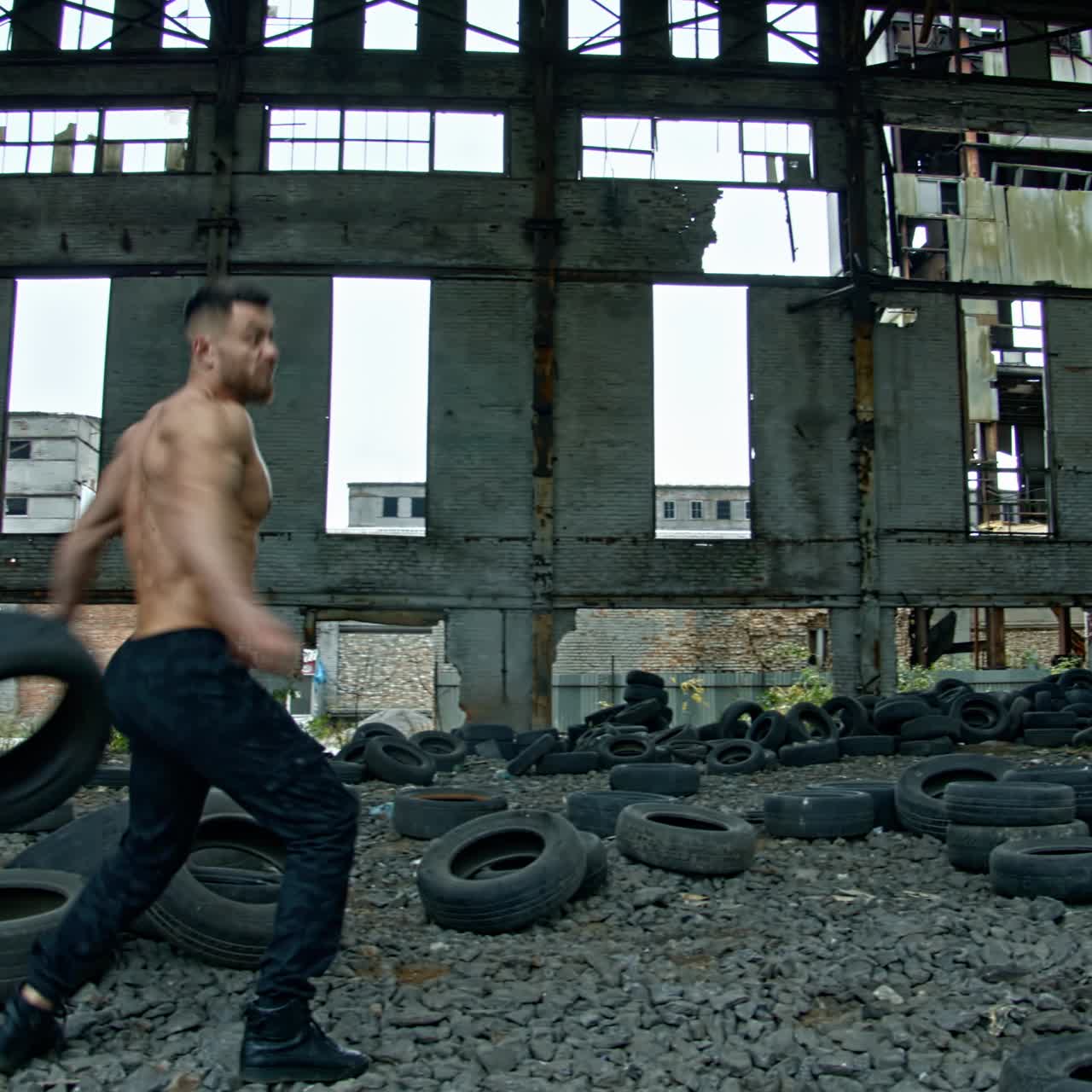 Shirtless man training in ruined plant. Strong athlete throwing old car tires while doing his workout in abandoned place. Cross fitness routine.