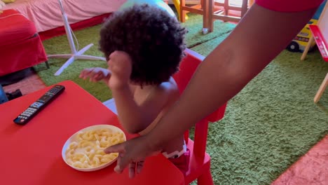 Exotic-and-playful-two-year-old-african-european-kid-eating-cheese-snacks-seated-in-a-plastic-red-table-at-home-in-a-hot-summer-day