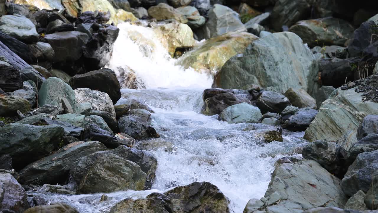Clear mountain stream water rushes over large mossy rocks in natural daylight. The camera remains steady, capturing dynamic water movement and textured stones