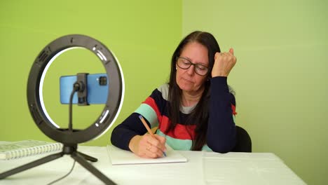 mujer madura contemplando mientras escribe en el bloc de notas durante una reunión virtual en la casa