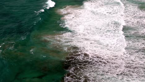 Toma-Aérea-De-La-Cresta-Espumosa-De-Una-Ola-Rompiendo-A-Lo-Largo-De-La-Costa,-La-Espuma-Del-Mar,-Parecida-A-Un-Encaje,-Encontrándose-Con-La-Playa.