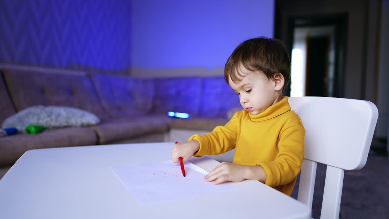 Charming little baby sitting at the desk drawing. Adorable child is focused on the hobby. Blurred backdrop.