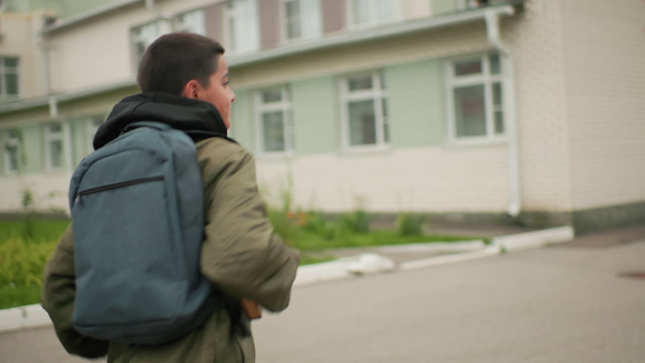 한 남자아이가 교실을 향해 달려가면서 팔 아래에 노트북을 끼고 어깨에는 백팩을 멘 채 학교 운동장에서 매일 학업에 집중하는 모습을 보여주고 있어요. 건물 외부 근처에서요.