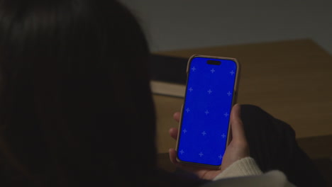 Over The Shoulder Shot Of Woman At Home Sitting On Sofa With Blue Screen Mobile Phone Scrolling Through Internet Or Social Media 3