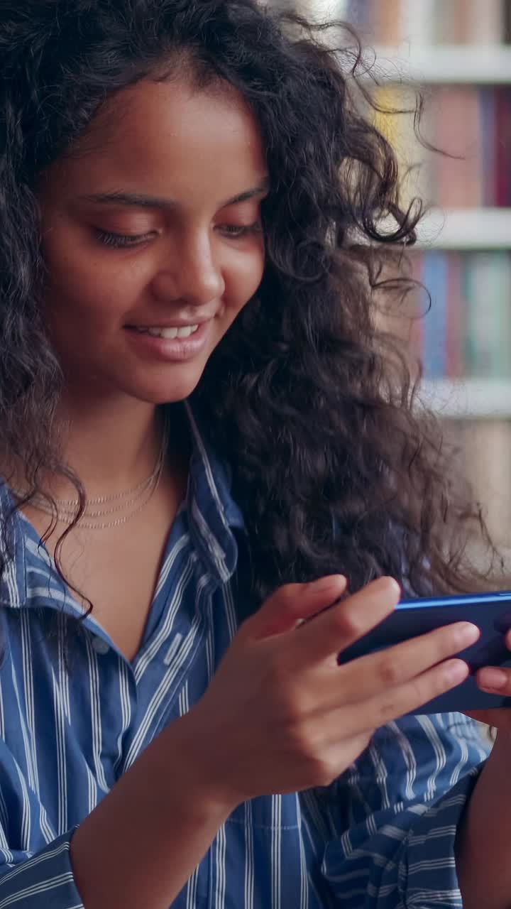 Young optimistic curly indian woman teen plays video games on smartphone
