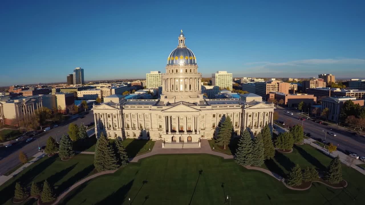 Aerial view of a grand government building with a golden dome, surrounded by lush greenery and urban skyline, showcasing architectural beauty and city life progression