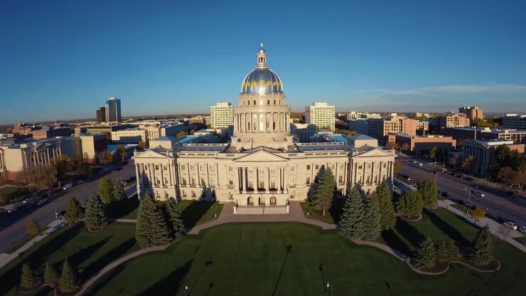 Vista aerea di un imponente edificio governativo con una cupola dorata, circondato da una vegetazione lussureggiante e dallo skyline urbano, che mette in mostra la bellezza architettonica e la progressione della vita cittadina