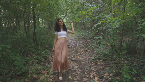 elegante giovane donna che cammina da sola nel sentiero forestale del parco naturale, carrello che segue il colpo esterno di una ragazza sottile e attraente che cammina toccando il ramo di un albero, modello brunetta alla moda casual