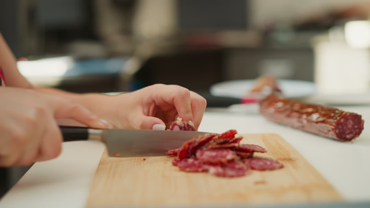 Confectioner hands slicing cured sausage into uniform thin rounds with sharp kitchen knife on wooden board over bright white table, showcasing precise technique and rich marbled meat texture