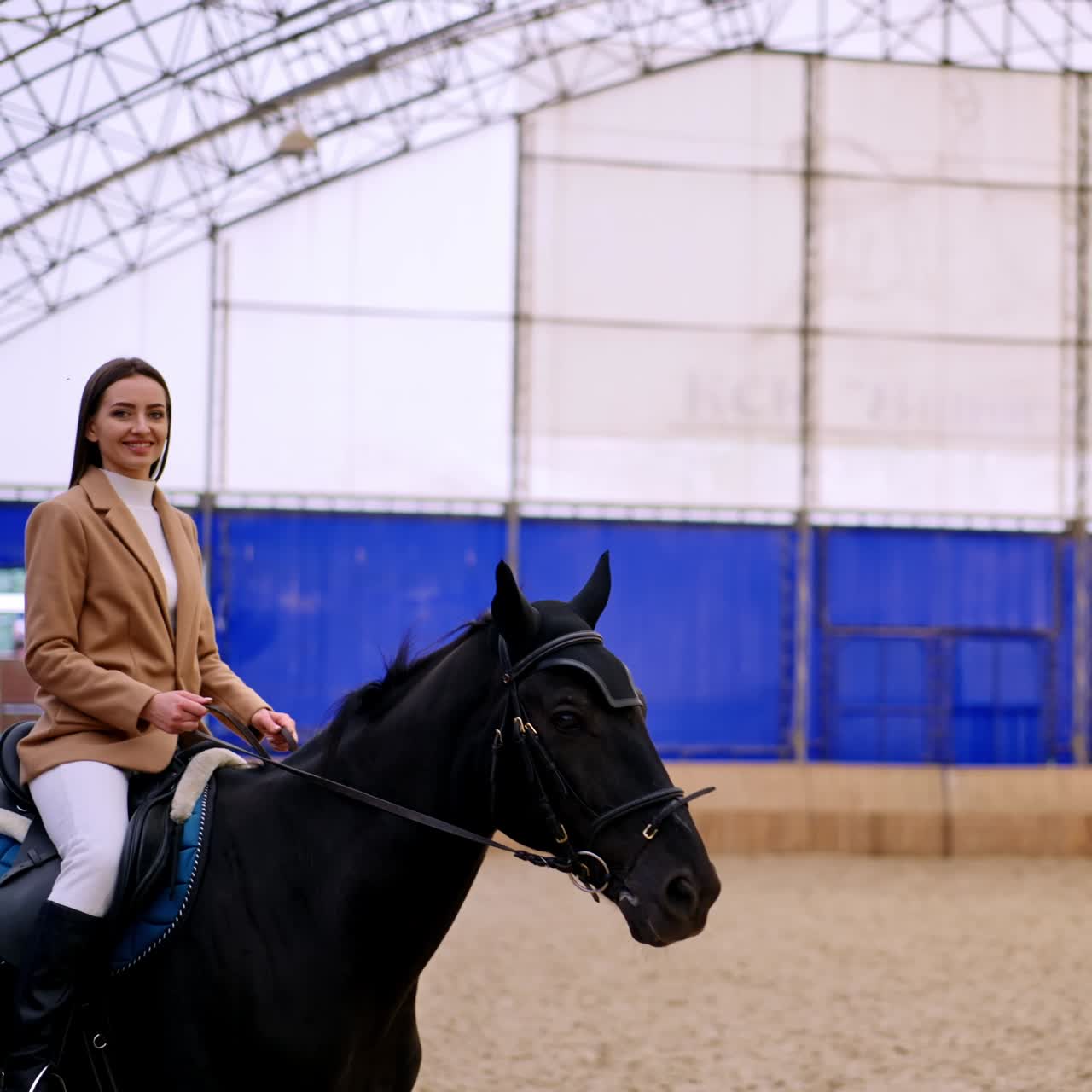 Happy smiling lady taking lessons of horse riding. Beautiful long-haired brunette rides on a black horse slowly. Manege backdrop