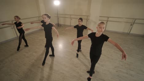 a group of young ballet students in black dancewear practicing positions in a spacious ballet studio with wooden flooring and wall-mounted barres. focused expressions and synchronized movements.