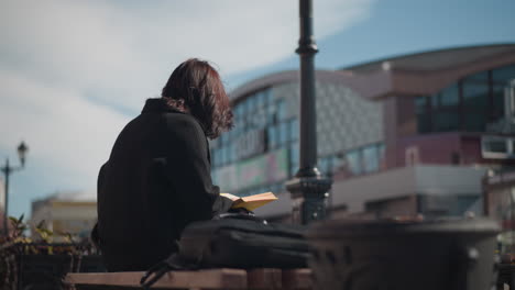 woman reading outdoor with her hair covering her face, turning a page, flower pots sway in the wind, while blurred background features urban store and lamp pole under a sunny sky