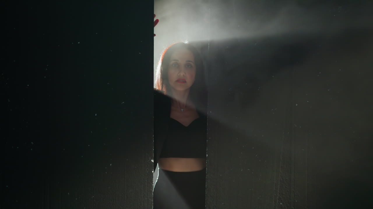 Mysterious woman in a black outfit emerges from the shadows, her hands pressed against a wall with a dramatic light illuminating her face as the camera zooms in