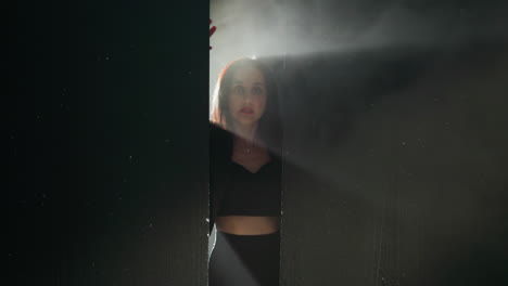 mysterious woman in a black outfit emerges from the shadows, her hands pressed against a wall with a dramatic light illuminating her face as the camera zooms in