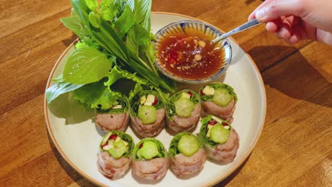 a hand drizzles sauce over vietnamese pork rolls on a wooden table. fresh herbs accompany the dish, enhancing its vibrant presentation