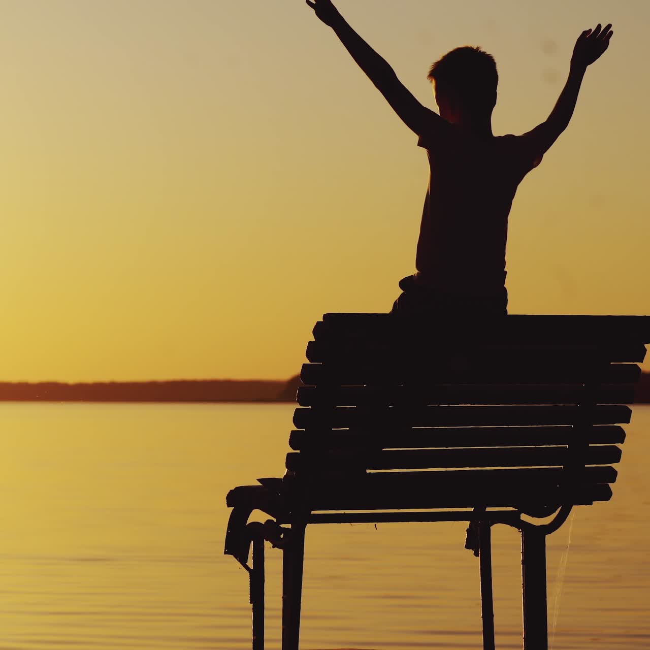 A little boy is looking at the sunset and wonderful landscape near the river on the bench. A guy raises his hands up. Weekend.