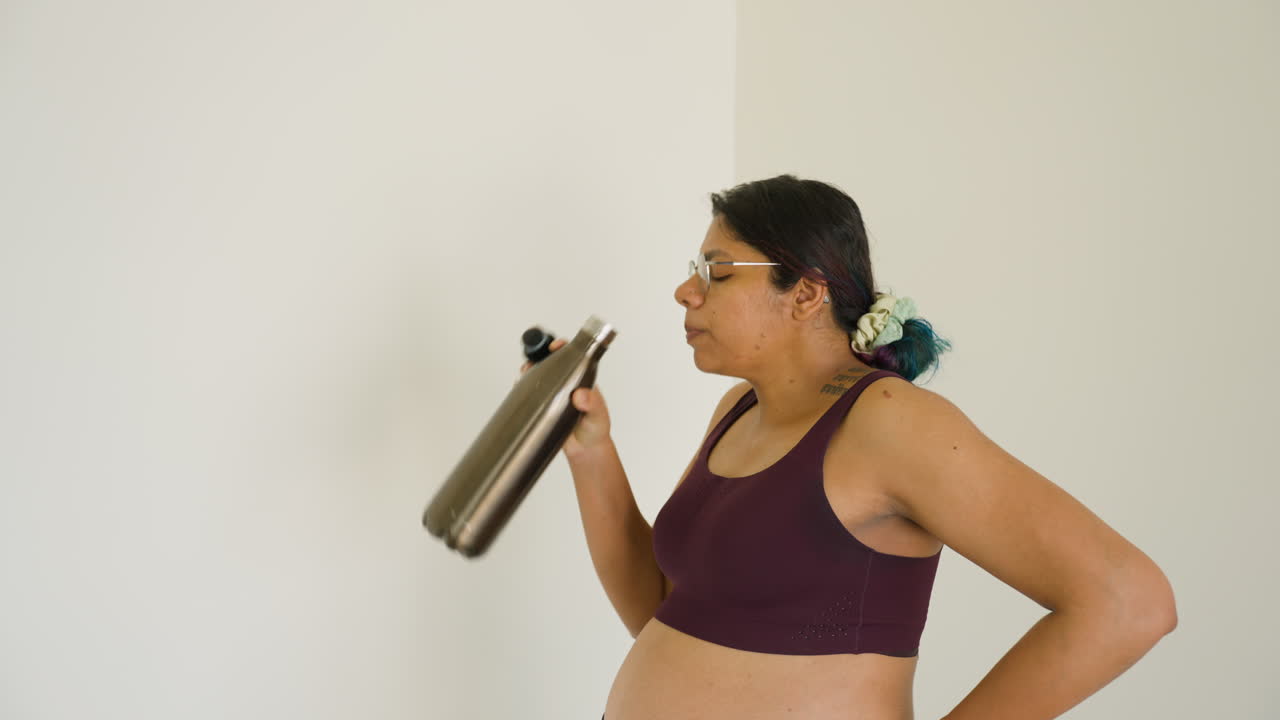 Giovane donna indiana al terzo trimestre che beve acqua per idratarsi dopo un allenamento di yoga