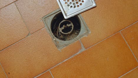 Dirty-old-water-floor-drain-with-opened-strainer,-brown-tiles-flooring