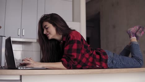 a girl in her 20's lying on kitchen table surface and typing something in laptop.