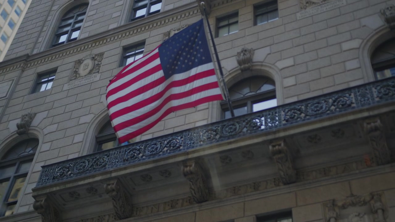 Bandiera americana pende da un balcone in ferro su una facciata in pietra scolpita con teste di leone, ghirlande e scudi araldici lavorati nella muratura, telecamera che si inclina verso l'alto all'interno di un taxi.