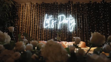 neon 'let's party' sign glowing on rustic wooden backdrop with white flowers in foreground