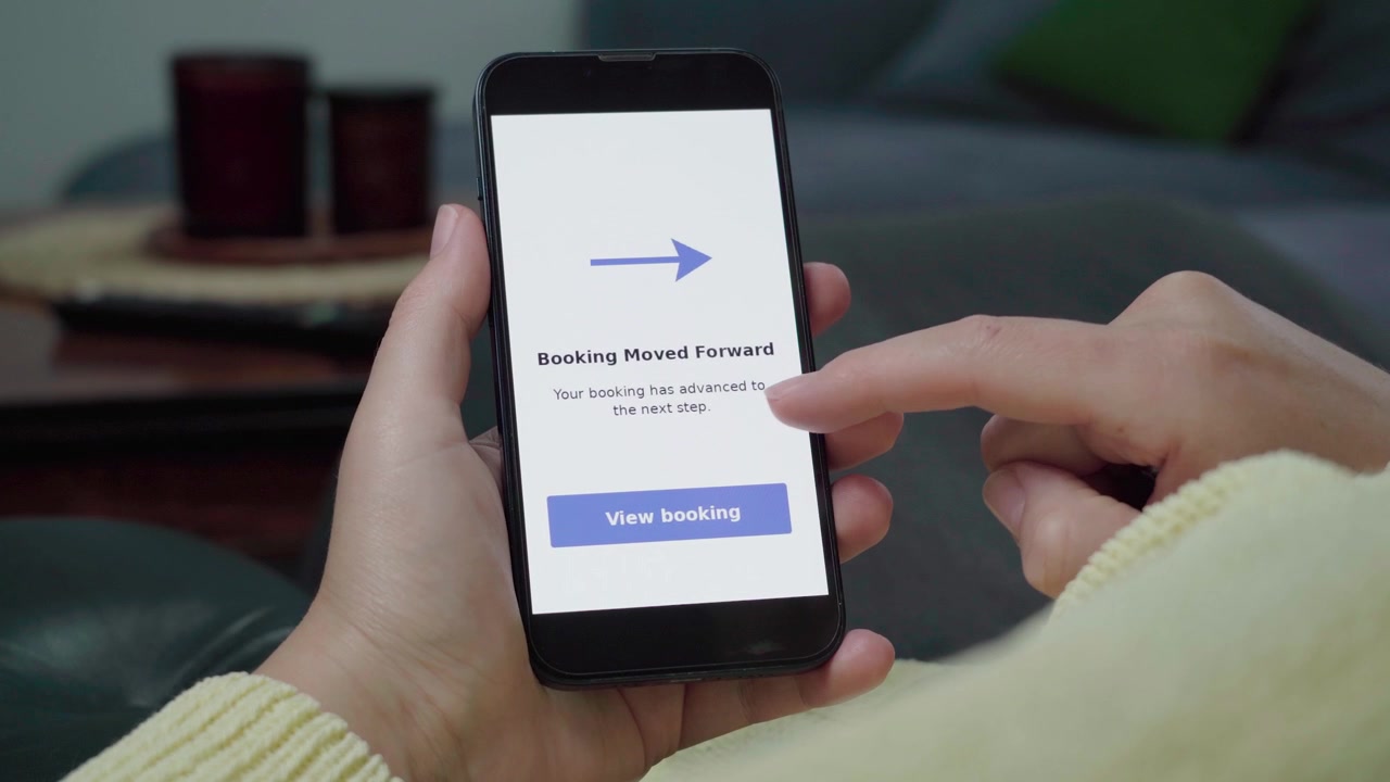 En person trykker på en mobilapp-skærm, der viser en notifikation om 'Booking flyttet frem' med en 'Se booking'-knap i en hyggelig stue.