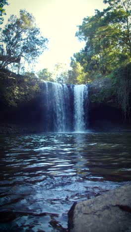 a pristine waterfall cascades from a rocky cliff into a serene pool below. lush green foliage frames the scene, sunlight dappling through the trees (vertical)