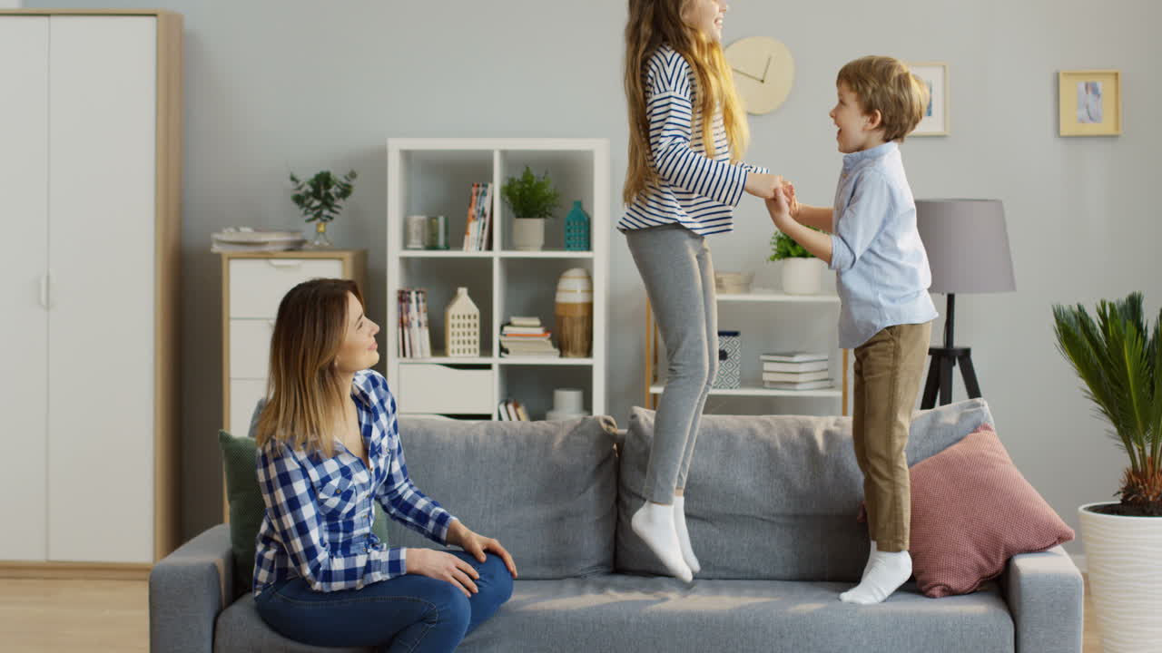Small Cute Children, Brother And Sister, Having Fun In The Living Room While Jumping On The Sofa And Their Mother Watching Them