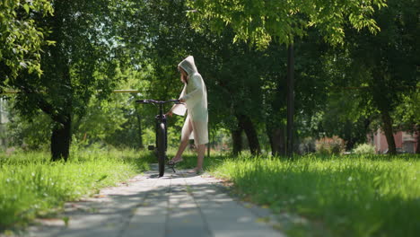 young woman in translucent raincoat strolls on sunny path with bicycle, stops to place stand, removes hood, and leans on handlebars,background includes lush greenery and shaded trees