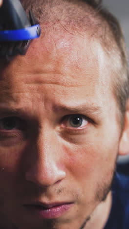 closeup portrait of young uncut bearded guy in front of mirror, cuts his hair head with clipper by himself, frowns, gets angry, focused