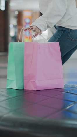 mujer sentada en un asiento de cuero en un centro comercial recoge bolsas de compras de menta y rosa antes de irse, con un fondo de venta al por menor borroso con tiendas y iluminación brillante