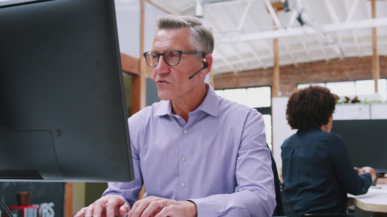 Mature Male Customer Services Agent Working At Desk In Call Center