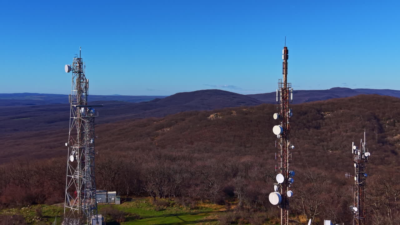 Communication towers stand tall on a high hill surrounded by trees under a clear blue sky during daylight hours in an open landscape