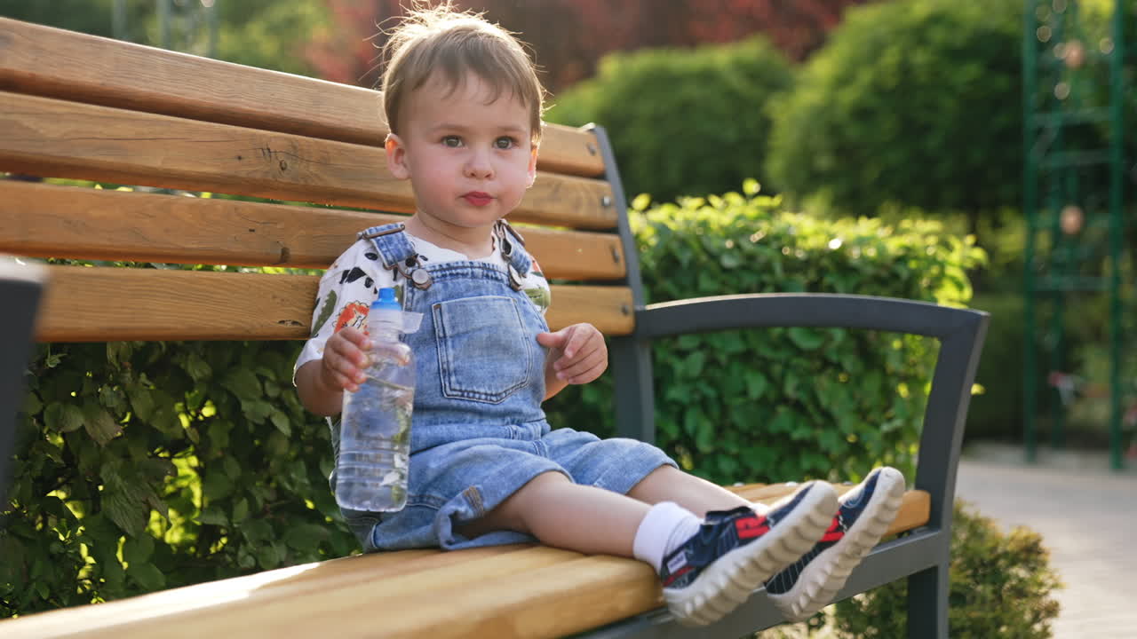 Smart little kid sits on the bench outdoors. Toddler boy drinks water from a bottle waving his feet.