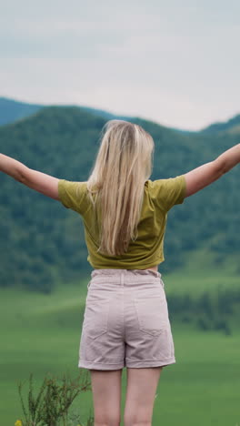 graciosa mujer rubia excursionista con las manos levantadas disfruta de la vista verde del valle de la montaña en la reserva ecológica salvaje en un día nublado vista trasera en cámara lenta