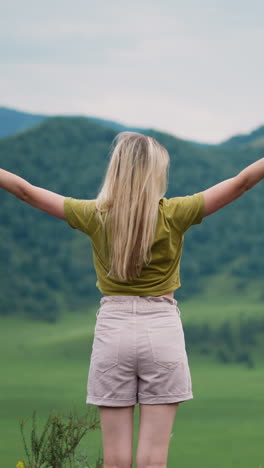 graceful blonde woman hiker with raised hands enjoys green mountain valley view at wild eco preserve on overcast day backside view slow motion