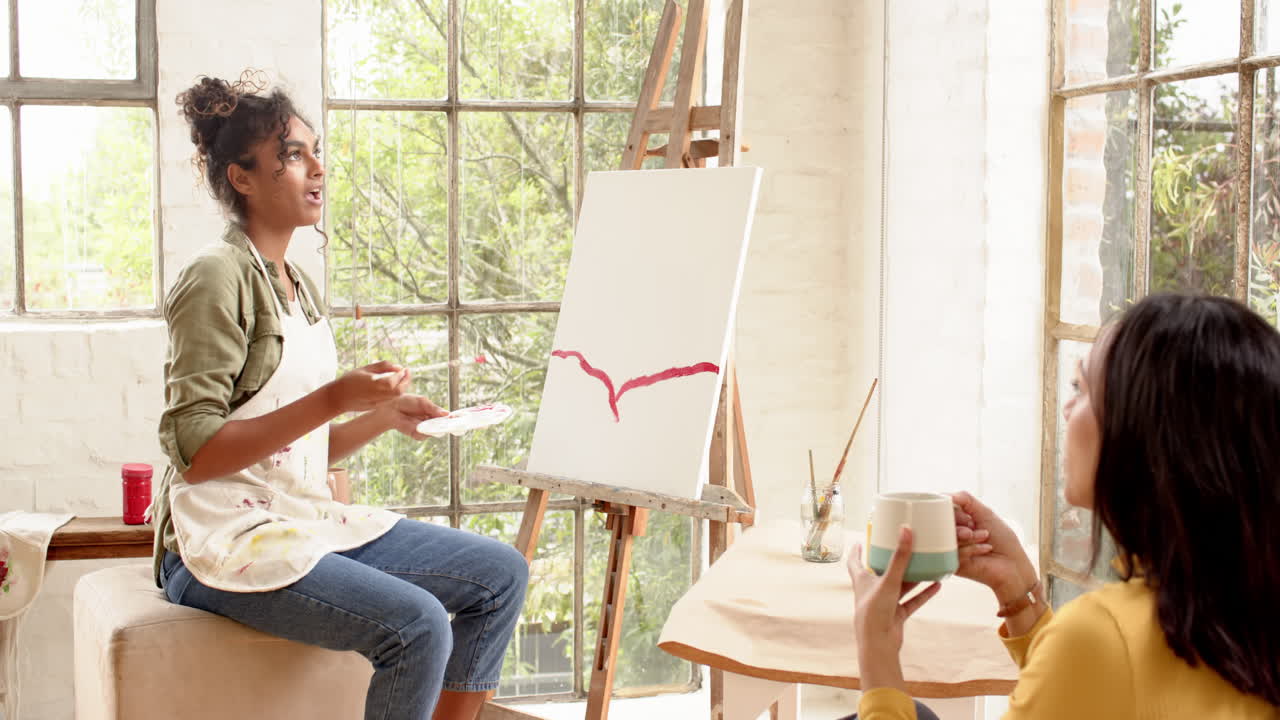 Female artist painting on canvas while friend enjoys coffee in bright home studio