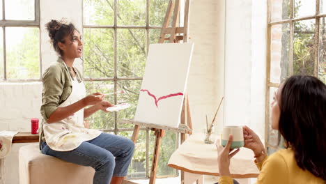 female artist painting on canvas while friend enjoys coffee in bright home studio