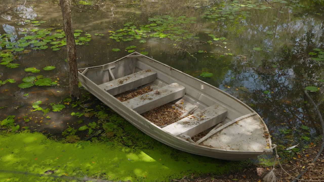 An old fishing tinny boat floats on the murky and mossy water of a lake, tied to the bank with trees shadowing the water and dried leaves filling the boat