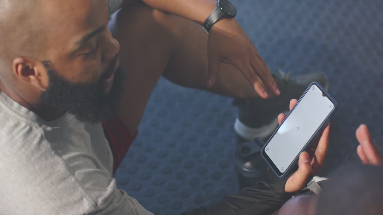 Adult African male sitting on gym floor holding phone, seeing loading icon, explaining to partner