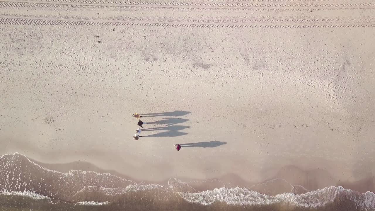 Drone shot from the sea to the shore, downward view. Camera following the movement of medium waves until it reaches group of people having a early walk on the beach.