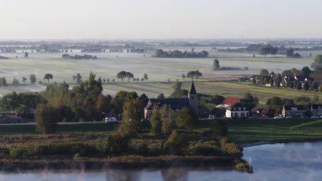 small riverside town with church tower in netherlands during rising fog, aerial view