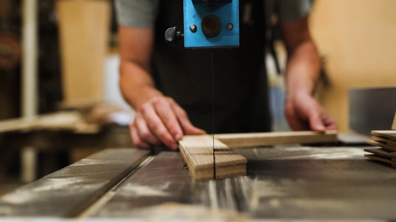 A skilled craftsperson focuses on making accurate cuts using a band saw in a well-lit woodworking shop. The workshop is filled with tools and wood pieces, reflecting a creative environment.