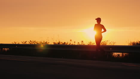 Silhouette-Of-A-Young-Active-Woman-Jogging-Along-A-Highway-Near-The-Sea-At-Sunset