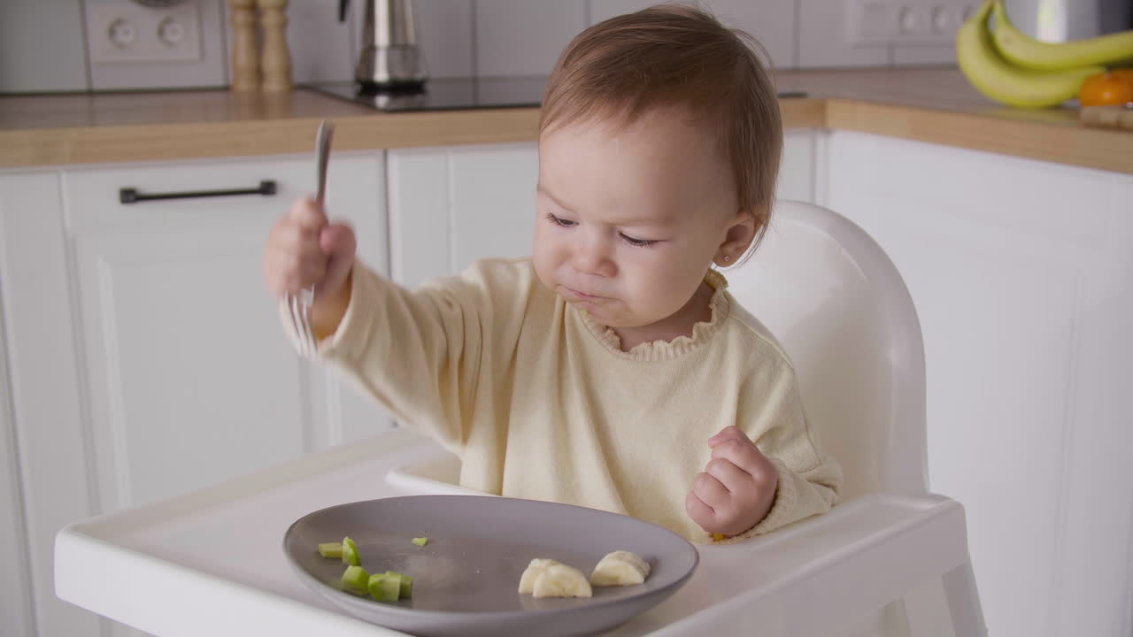 Søt babyjente sitter i høystol på kjøkkenet og holder en gaffel og spiser fruktskiver.