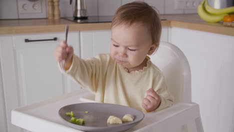 linda niña sentada en una silla alta en la cocina y sosteniendo un tenedor y comiendo rodajas de fruta