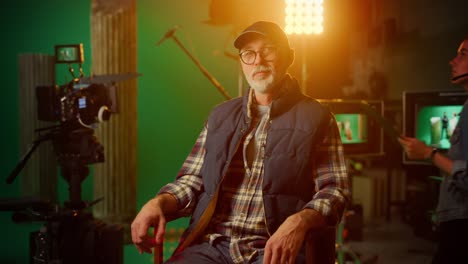 protrait of prominent successful senior director sitting in a chair on a break looking at camera cheerfully. on the studio film set with high end equipment professional crew shooting high budget movie